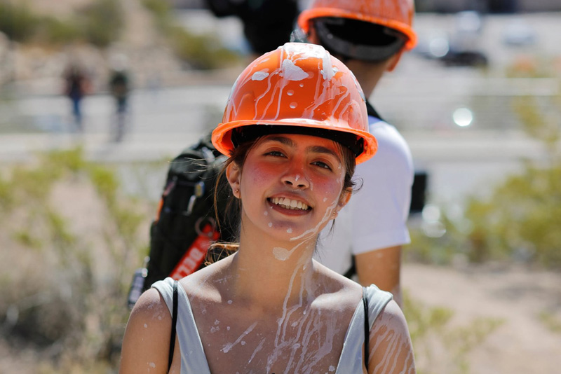 Close-up of a participant wearing an orange hard hat splattered with white paint, with paint also visible on the face and clothing, standing outdoors in bright sunlight. Another participant with a backpack stands in the background on a rocky hillside near a paved area.