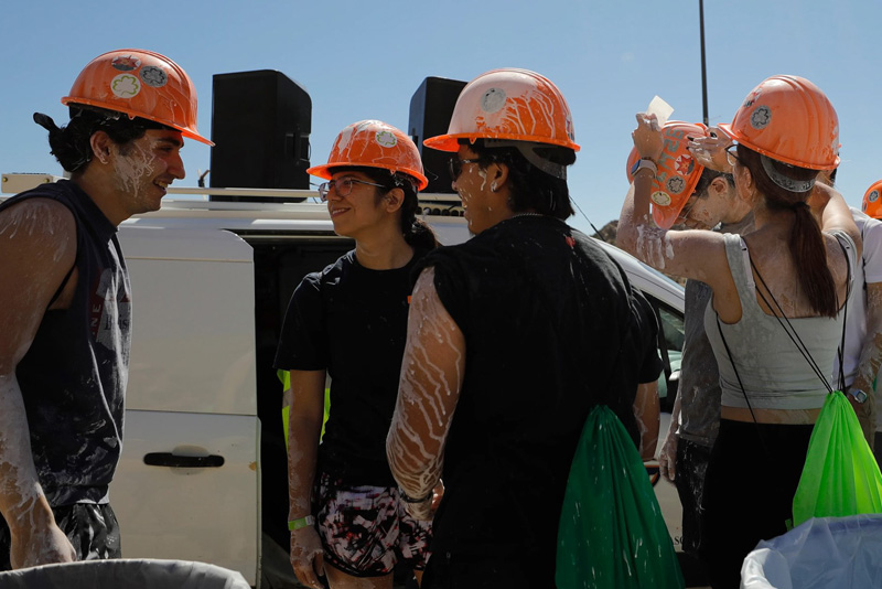 Participants wearing orange hard hats, splattered with white paint, stand and talk near a white utility vehicle on a sunny hillside. Several individuals adjust their helmets while others face each other, with green drawstring bags visible and a clear blue sky in the background.