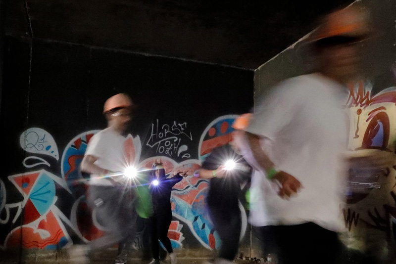 A group of participants wearing orange hard hats moves quickly through a dimly lit underpass covered in colorful graffiti, with motion blur capturing their movement. Several individuals hold bright lights that illuminate the walls and artwork along the tunnel.