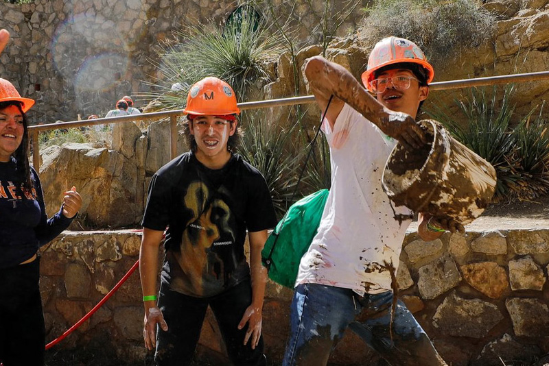 Participant wearing an orange hard hat lifts a muddy bucket and splashes its contents forward while standing in a muddy work area, with dirt covering arms and clothing. Other helmeted participants stand nearby against a stone wall and desert plants, all engaged in the outdoor activity under bright sunlight.