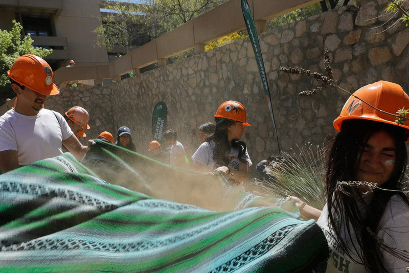 Participants wearing orange hard hats grip and shake a large green-and-black striped blanket, kicking up dust during an outdoor team activity. A stone wall, desert plants, and additional participants are visible in the background under bright sunlight.
