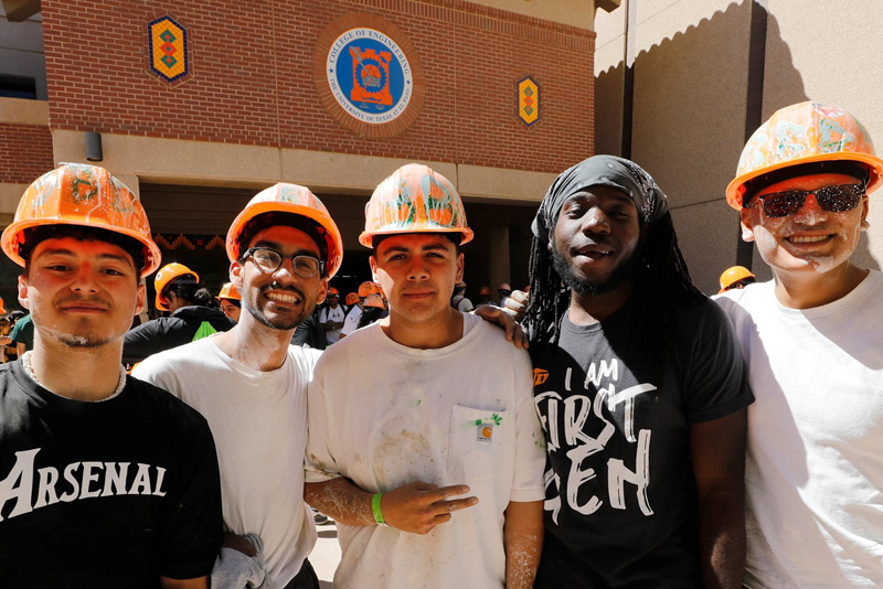 Five participants wearing orange hard hats stand together in front of a brick university building, posing for a group photo during an outdoor event. The building behind them features a circular emblem and colorful tile accents, while paint splatters are visible on the participants’ helmets and clothing.