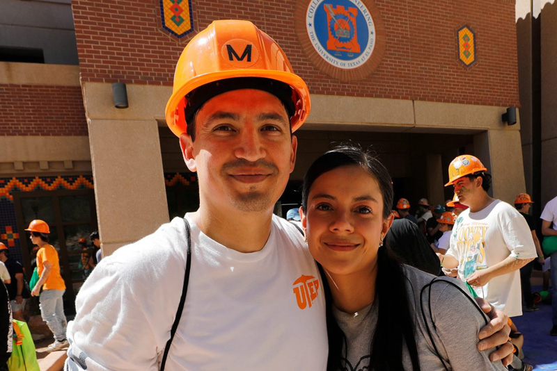 Two participants wearing orange hard hats stand together in front of a brick campus building with a circular university emblem, posing closely for a photo. Other participants in helmets gather in the background, with colorful tile accents visible on the building under bright sunlight.