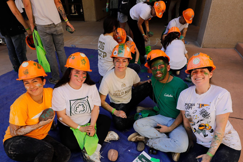 Five participants wearing orange hard hats sit together on the ground, posing for a group photo while surrounded by chalk markings and scattered rocks during an outdoor campus activity. Additional participants wearing helmets work in the background near a building entrance under bright daylight.