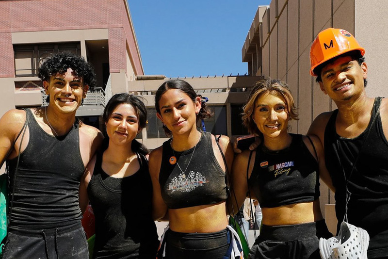 Five participants stand close together on a sunny campus walkway, posing for a group photo between modern university buildings. One participant wears an orange hard hat while others wear black athletic clothing, with clear blue sky and campus architecture in the background.