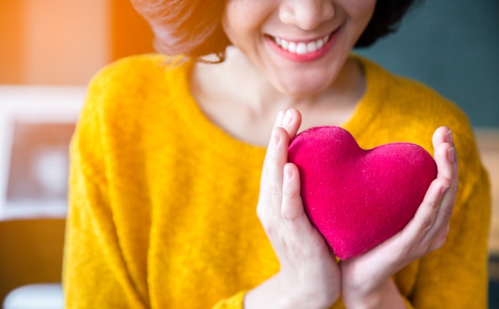A smiling woman in yellow sweater, holding a pink heart – How to Stay Healthy While Attending College Online | UTEP Connect