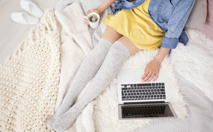 Woman in yellow dress, jean jacket, and gray stockings is sitting on bed with blankets, laptop, and coffee.