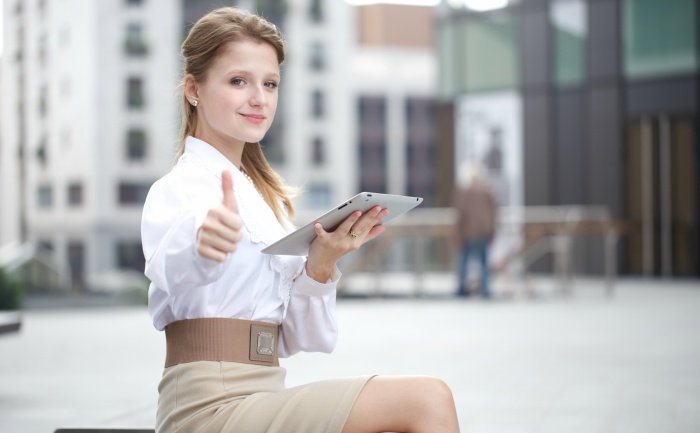 Young woman in professional wear holding a tablet, smiling, and having her thumb up as a sign of OK while sitting outside in an urban setting