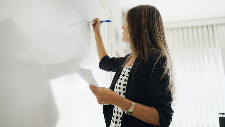 A woman stands next to a whiteboard in a classroom. The board displays English phrasal verHow to Become an ESL Teacher. Female teacher writing on a whiteboard while holding a worksheet during an ESL lesson in a bright classroom.bs with their Portuguese translations.