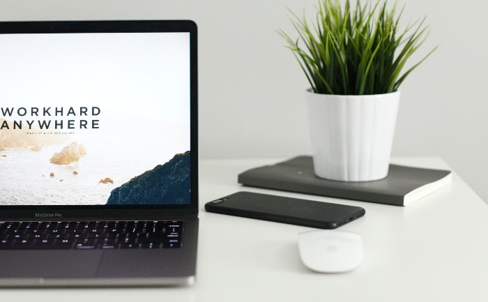 An open laptop with a screensaver that reads work hard anywhere next to a cell phone, notebook, and a plant, on a white desk table