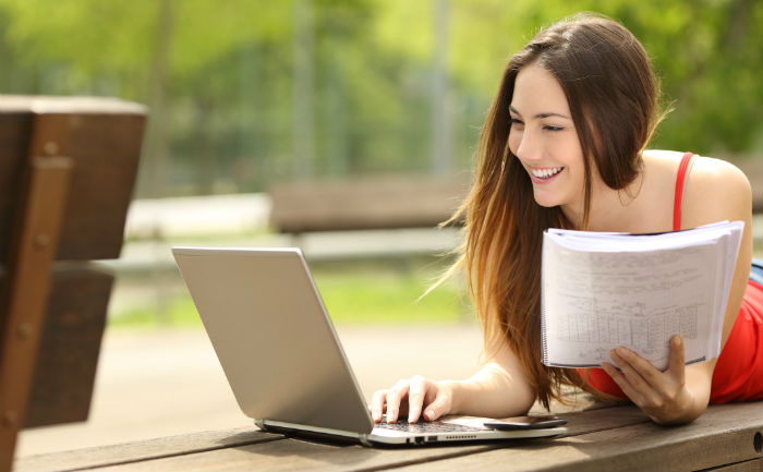 Woman holding a notebook and studying with a laptop on a bench outside.