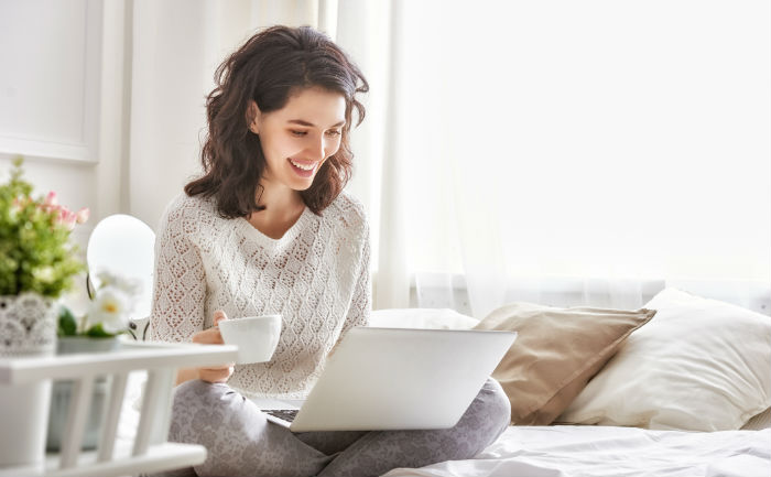 A woman in a white sweater holds her coffee mug while working on her online degree in a sunny room.