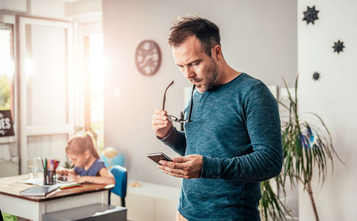 Man looks for online learning tips on his smartphone while his daughter works on homework at a desk.